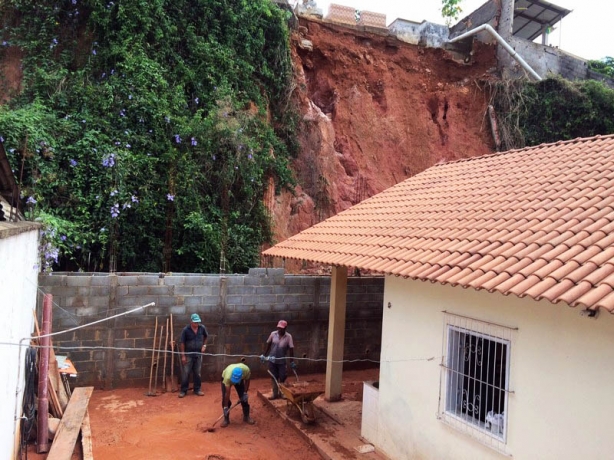 Acima v&ecirc;-se o barranco que desmoronou e parte dos t&uacute;mulos do cemit&eacute;rio. Na casa atingida, homens fazem a limpeza